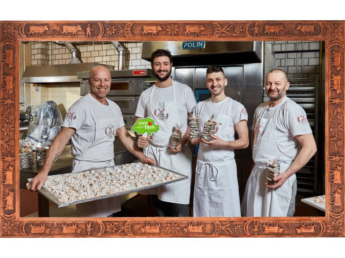 Vier Bäcker in einer Küche präsentieren Backwaren und verpackte Kekse, umrahmt von einem Holzrahmen.