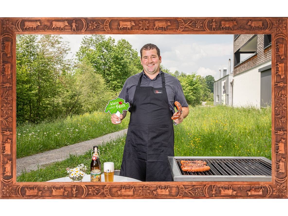 Mann grillt Würstchen, hält ein Schild mit „klein aber fein“ und ein Bier auf einem Tisch.