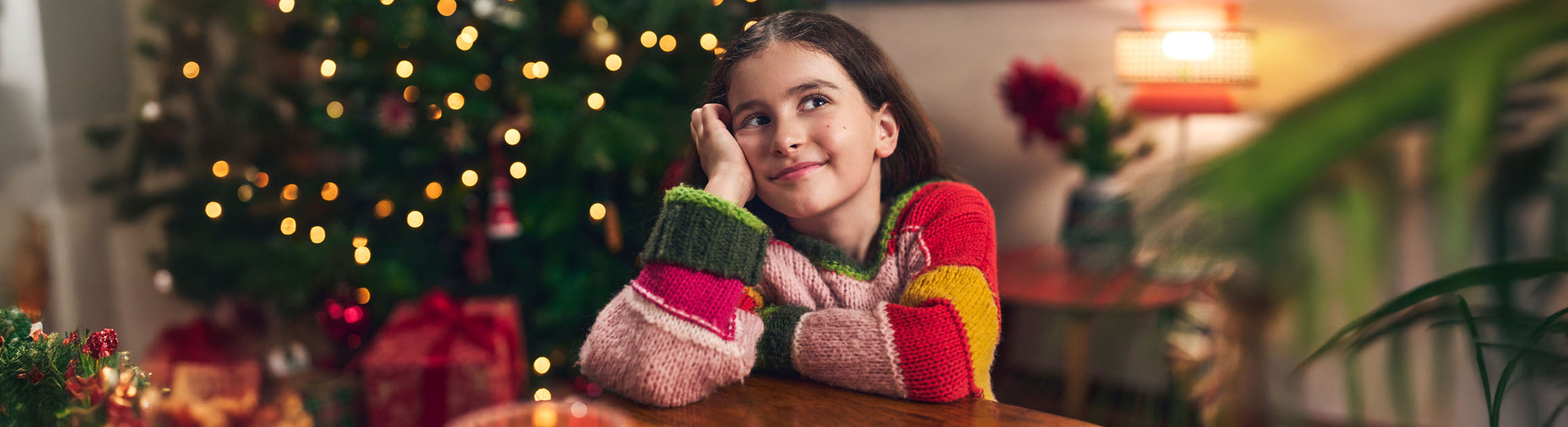 Fille souriante en pull coloré, appuyée sur une table avec un sapin de Noël en arrière-plan.
