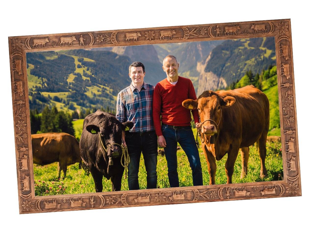 Zwei Männer mit Kühen auf einer Bergwiese, gerahmt von einem Holzschnitzrahmen.
