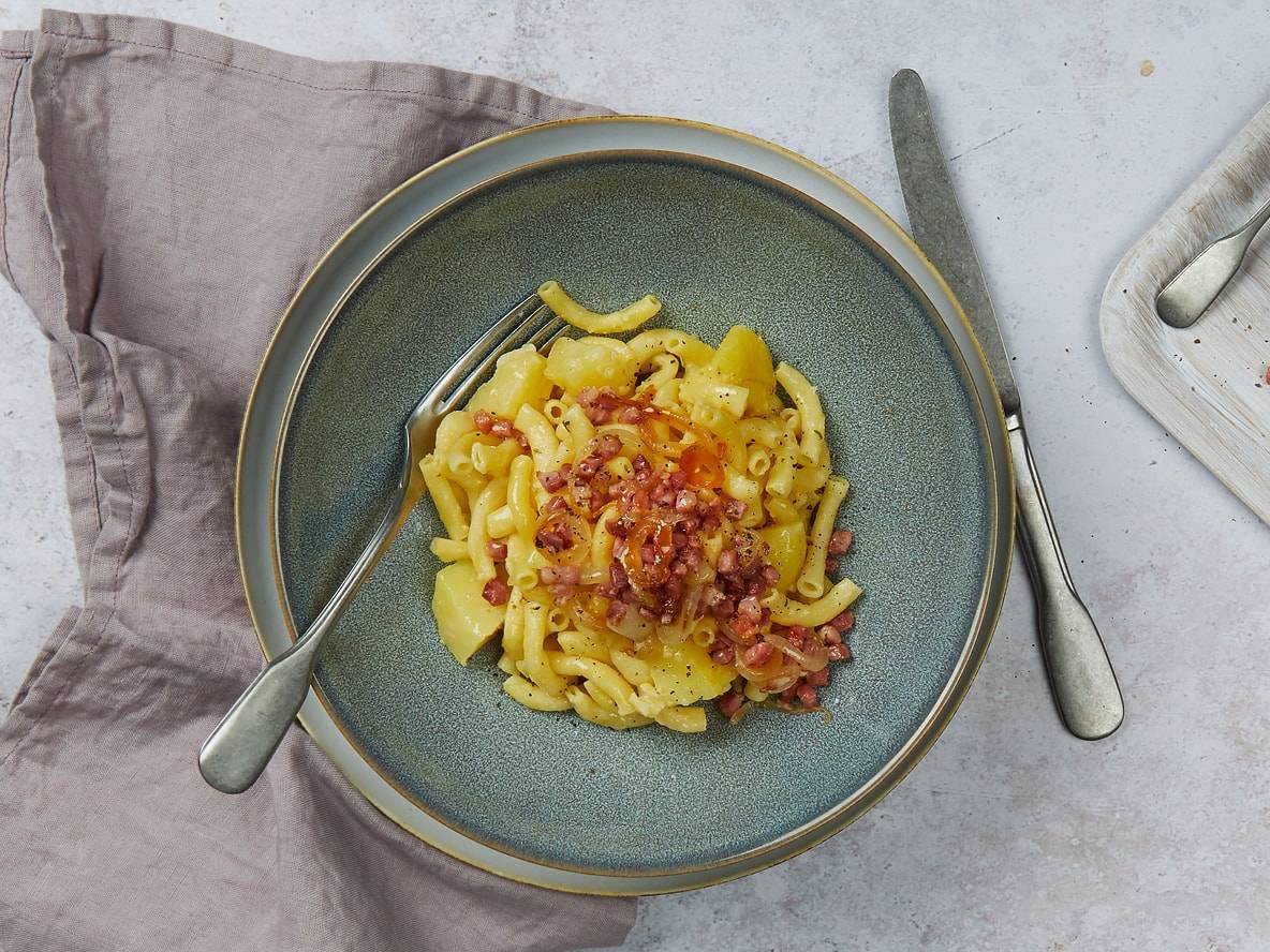 Macaronis aux pommes de terre et lardons croustillants dans un bol bleu, avec une fourchette et un couteau.