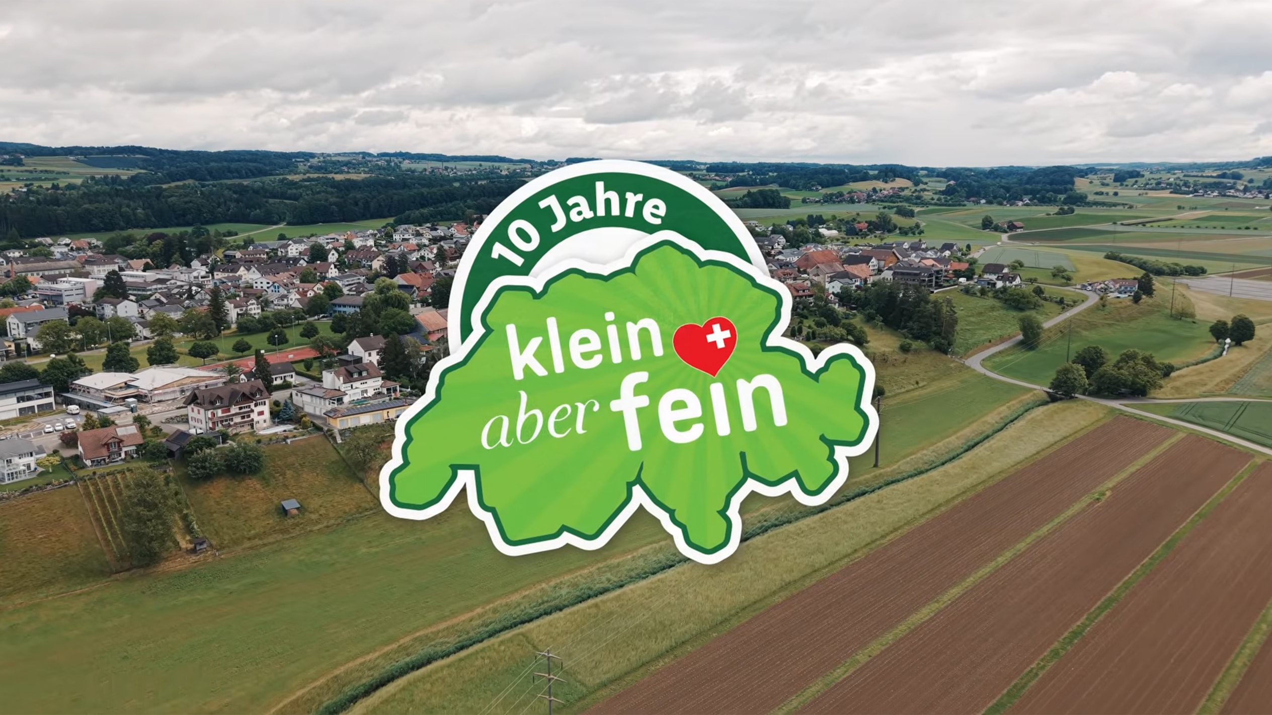 Luftaufnahme einer ländlichen Schweizer Landschaft mit einem Logo: '10 Jahre klein aber fein'.
