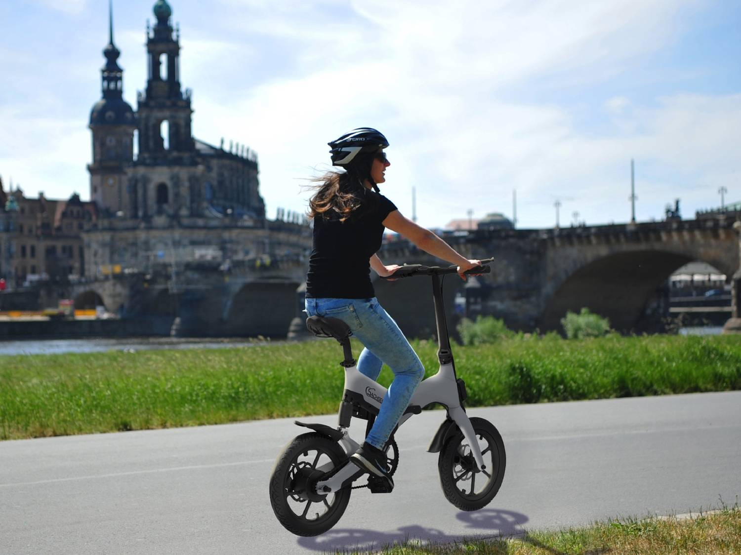 Donna su una bici elettrica pieghevole su una strada, con un ponte e un edificio storico sullo sfondo.