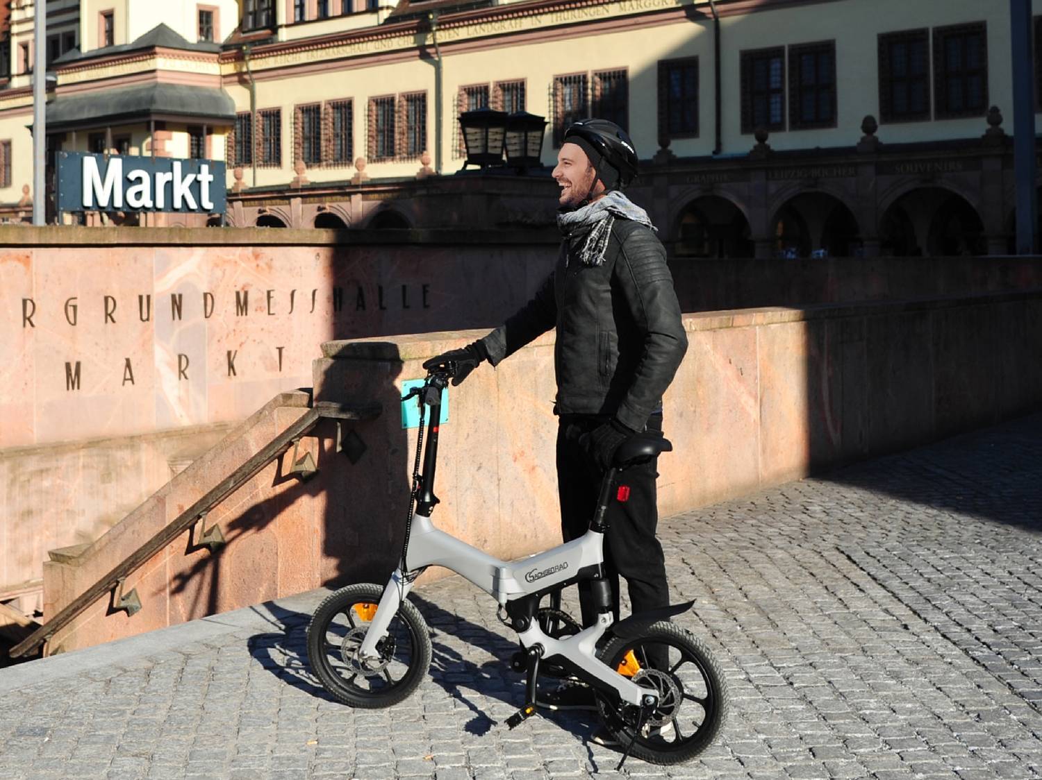 Un uomo sorridente con casco e sciarpa, in piedi accanto a una bici elettrica pieghevole grigia, davanti a un cartello 'Markt'.