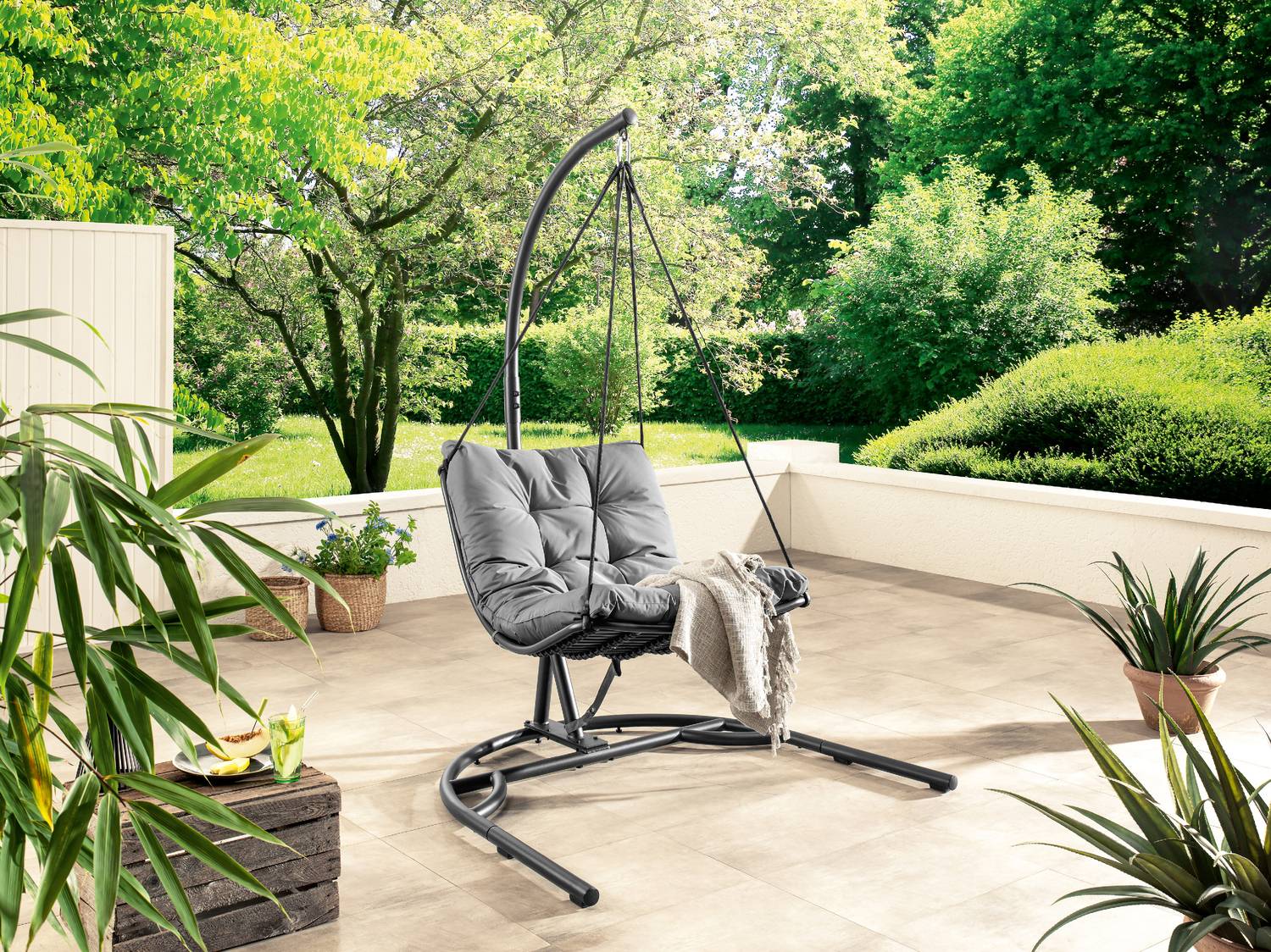 Poltrona sospesa grigia su una terrazza soleggiata con piante e un giardino verde.