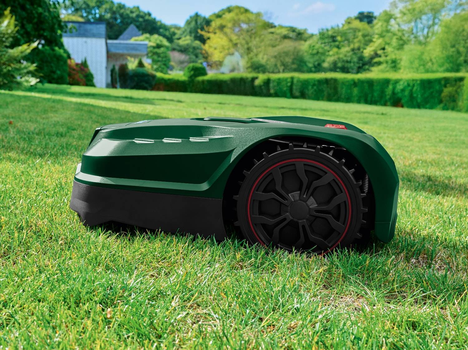 Grüner Mähroboter auf grünem Rasen mit Haus und Bäumen im Hintergrund.