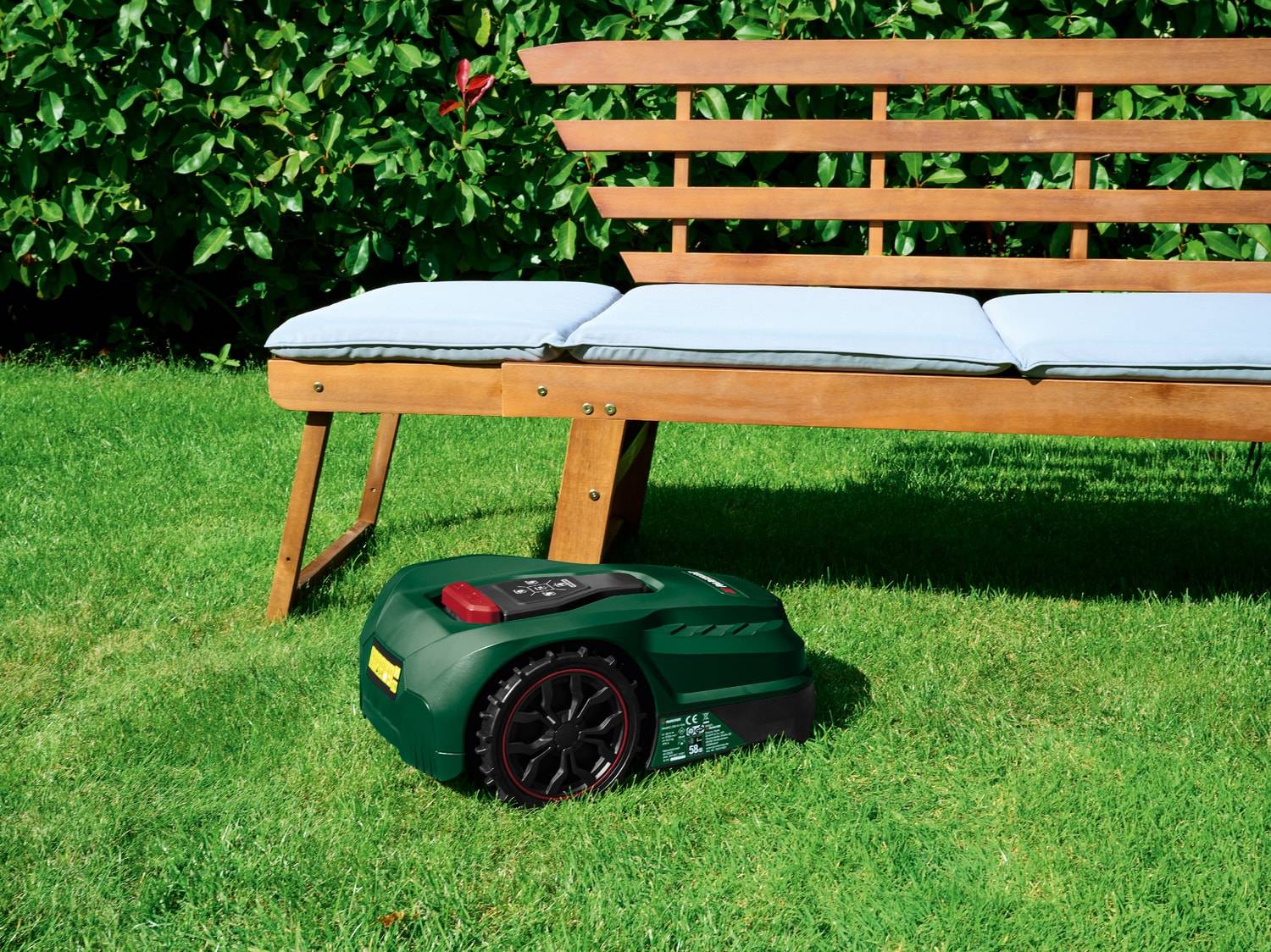 Grüner Mähroboter auf grünem Rasen mit Holzbank und hellen Kissen.