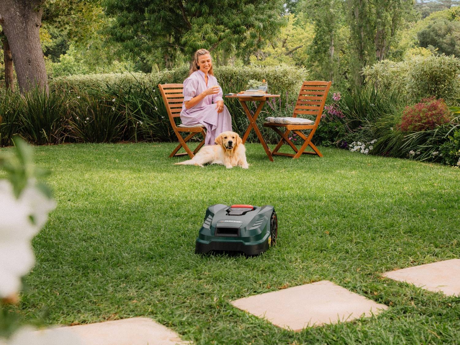 Ein Mähroboter auf dem Rasen, mit einer Frau und einem Hund, die im Hintergrund an einem Gartentisch sitzen.