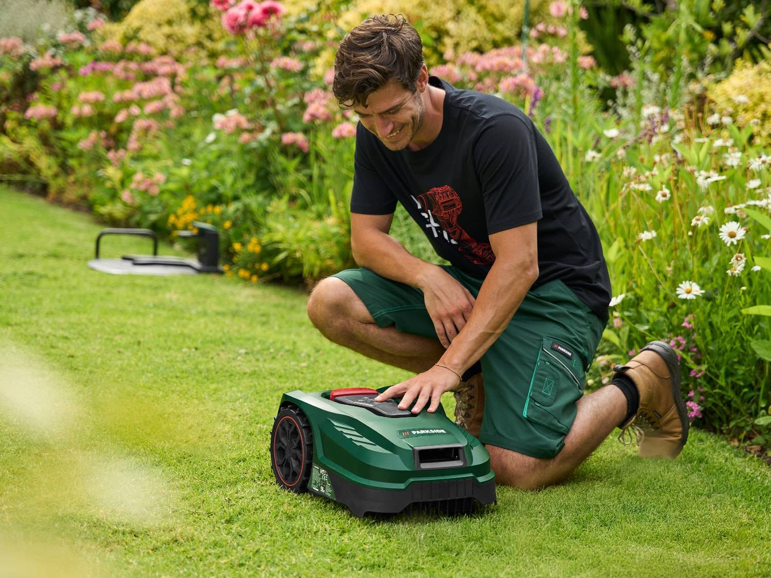 Lächelnder Mann hockt auf dem Rasen und stellt einen grünen Mähroboter in einem blühenden Garten ein.