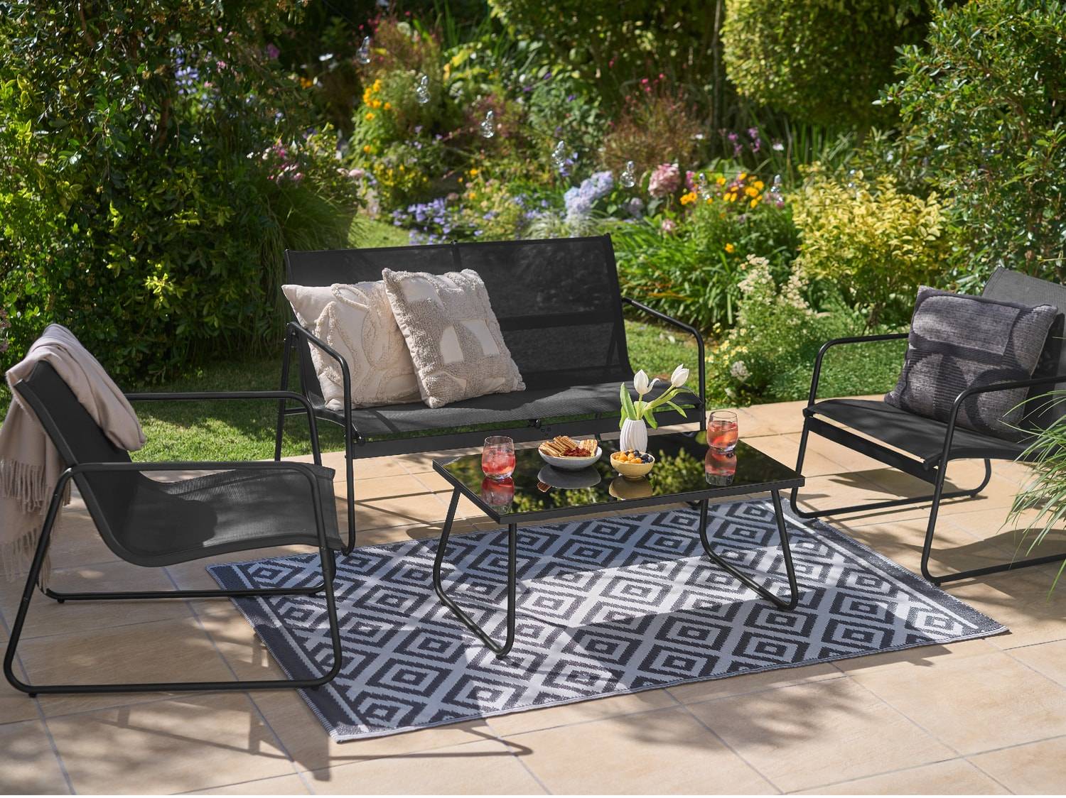 Schwarze Gartenmöbel mit Sofa, Sesseln, Couchtisch und geometrischem Teppich auf der Terrasse.