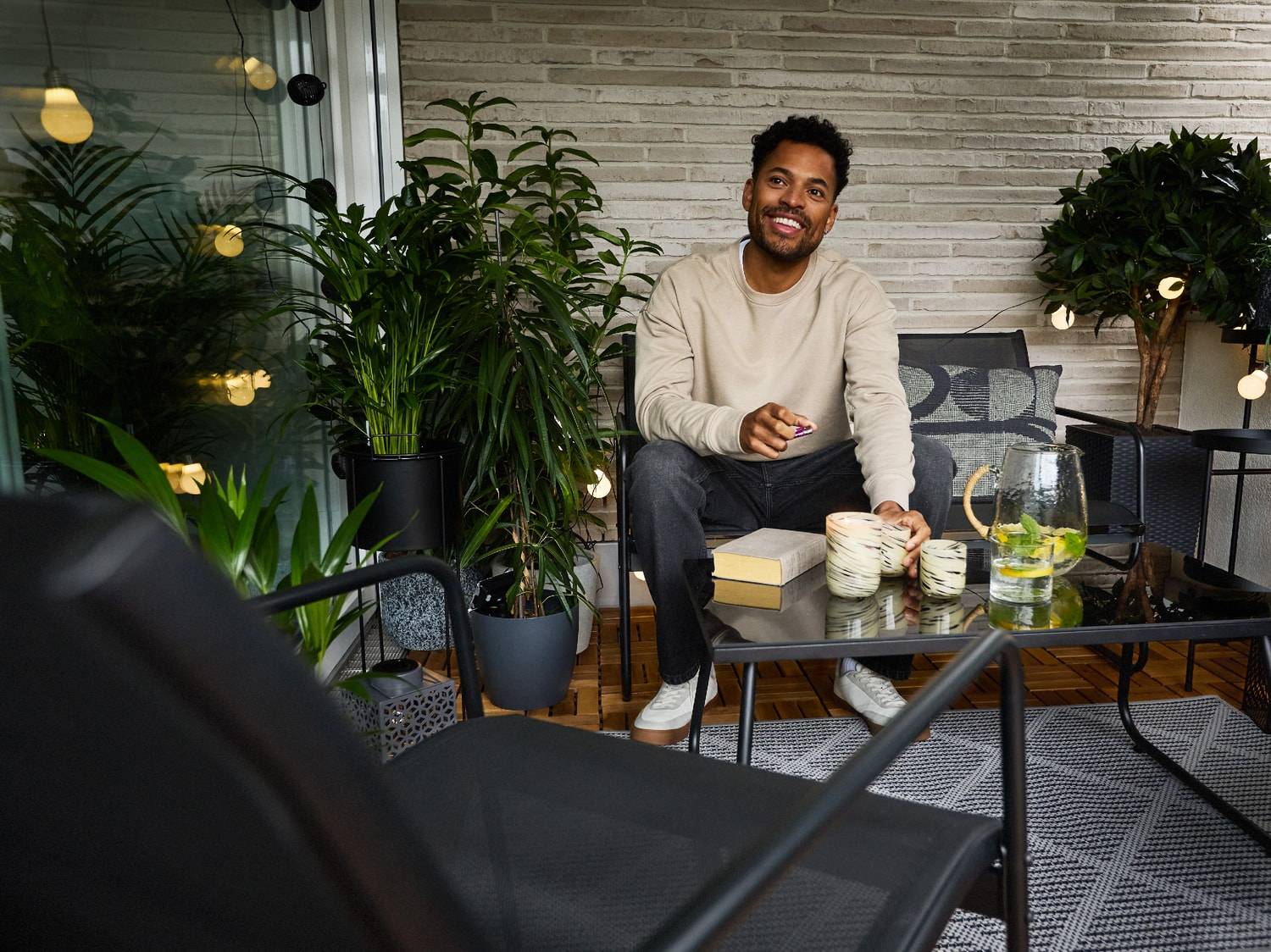 Lächelnder Mann sitzt auf einem Balkon mit Gartenmöbeln, Pflanzen und Lichterketten.
