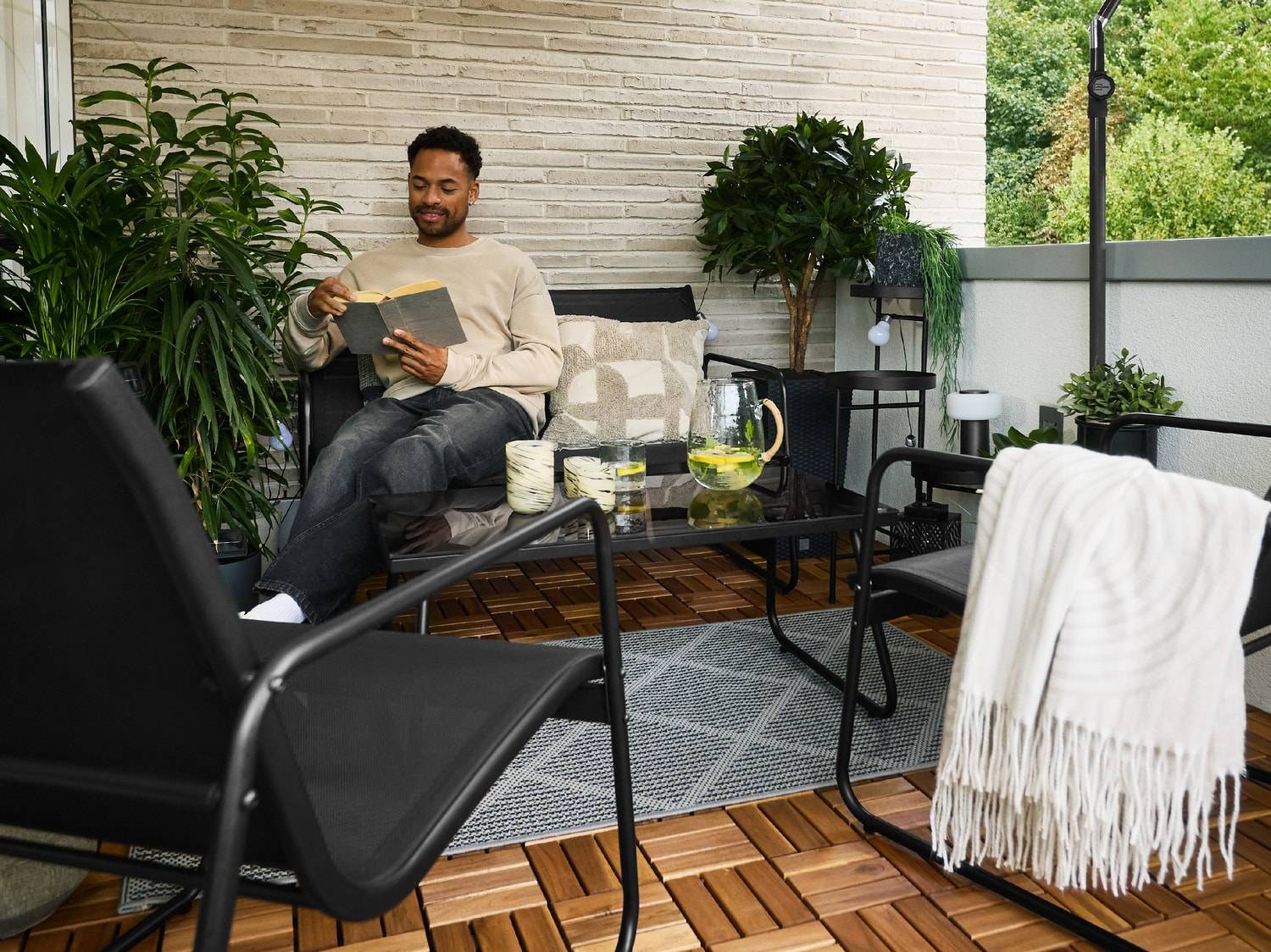 Mann liest auf einem Balkonsofa mit Tisch, Stühlen und Pflanzen.