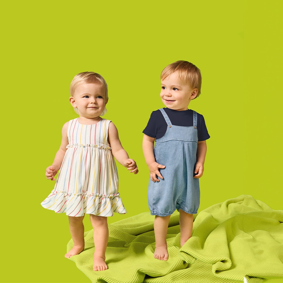 Zwei Kleinkinder, ein Mädchen im gestreiften Kleid und ein Junge im Jeans-Overall, stehen auf einer grünen Decke.