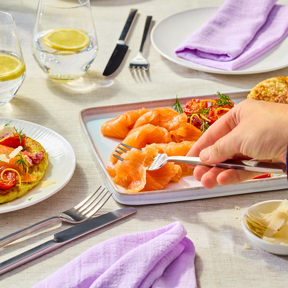 Eine Hand nimmt mit einer Gabel geräucherten Lachs von einer Platte auf einem gedeckten Tisch.