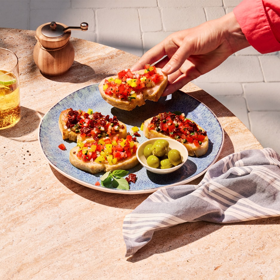Hand nimmt Bruschetta mit Tomaten- und Paprikabelag von blauem Teller mit Oliven.