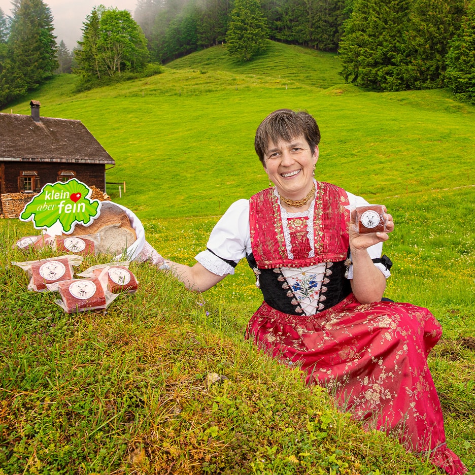 Frau in Tracht mit Brot und verpackten Fleischprodukten, im Hintergrund ein Bauernhaus.