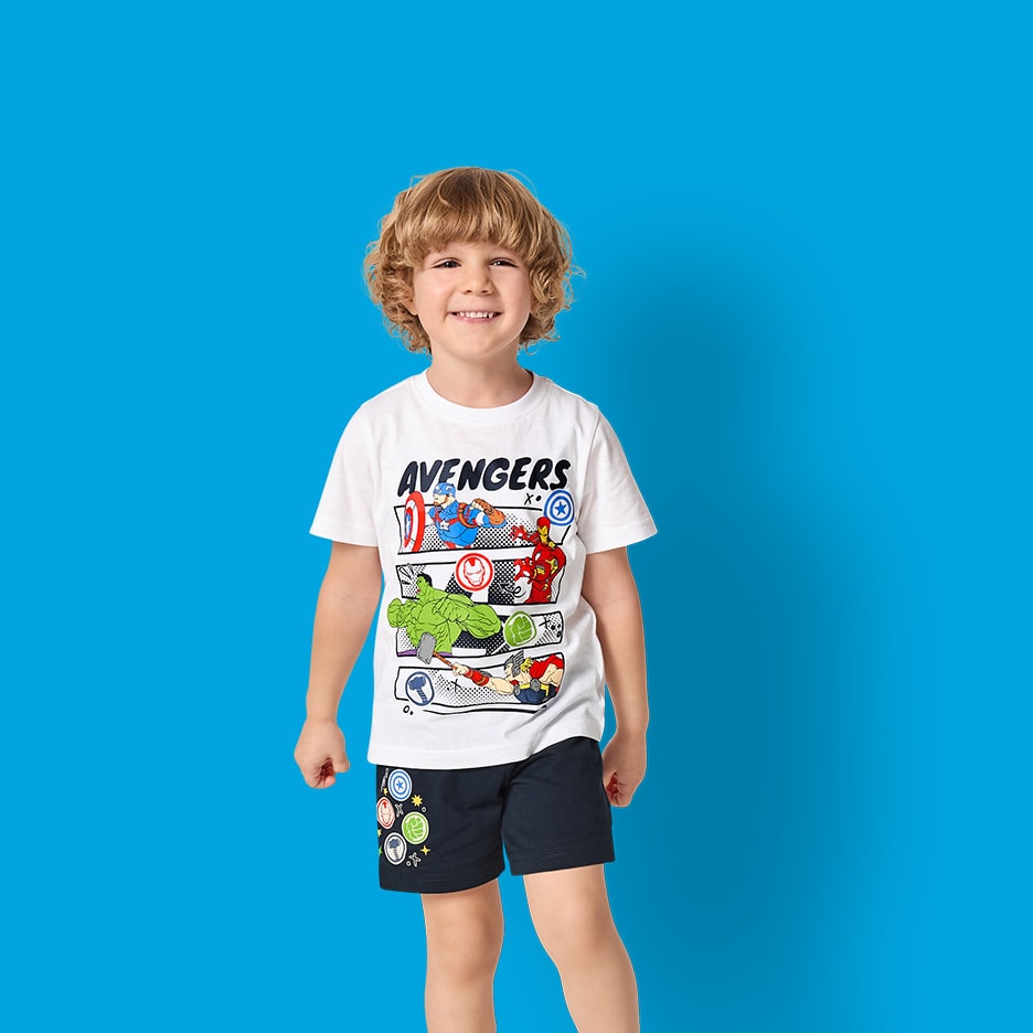 Kleiner Junge in weißem T-Shirt mit Superhelden-Aufdruck und marineblauen Shorts.