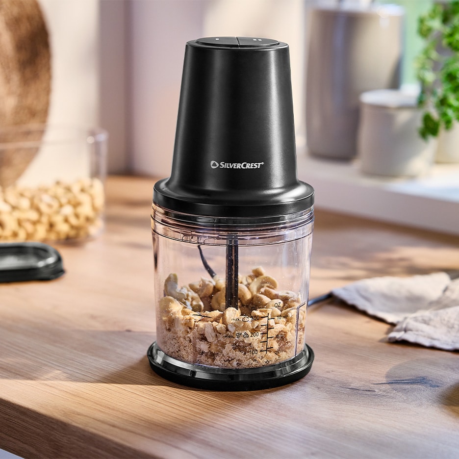 Ein schwarzer elektrischer Zerkleinerer mit gehackten Cashewnüssen auf einem Holztisch.