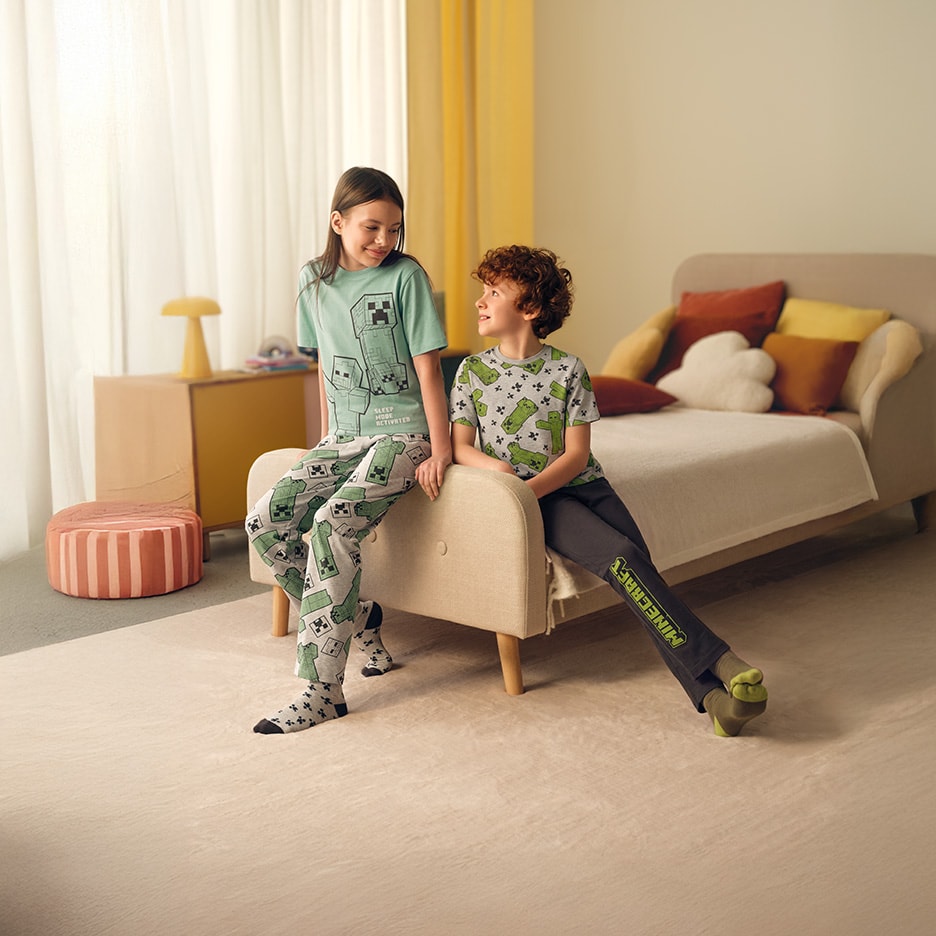 Zwei Kinder in Schlafanzügen mit Videospiel-Motiv, auf einem Sofa in einem Schlafzimmer sitzend.
