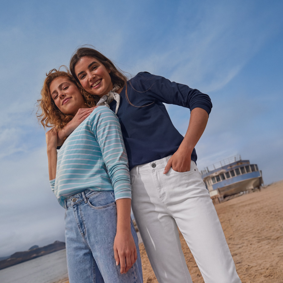 Zwei Frauen in Langarmshirts und Jeans an einem Strand mit einem Boot.