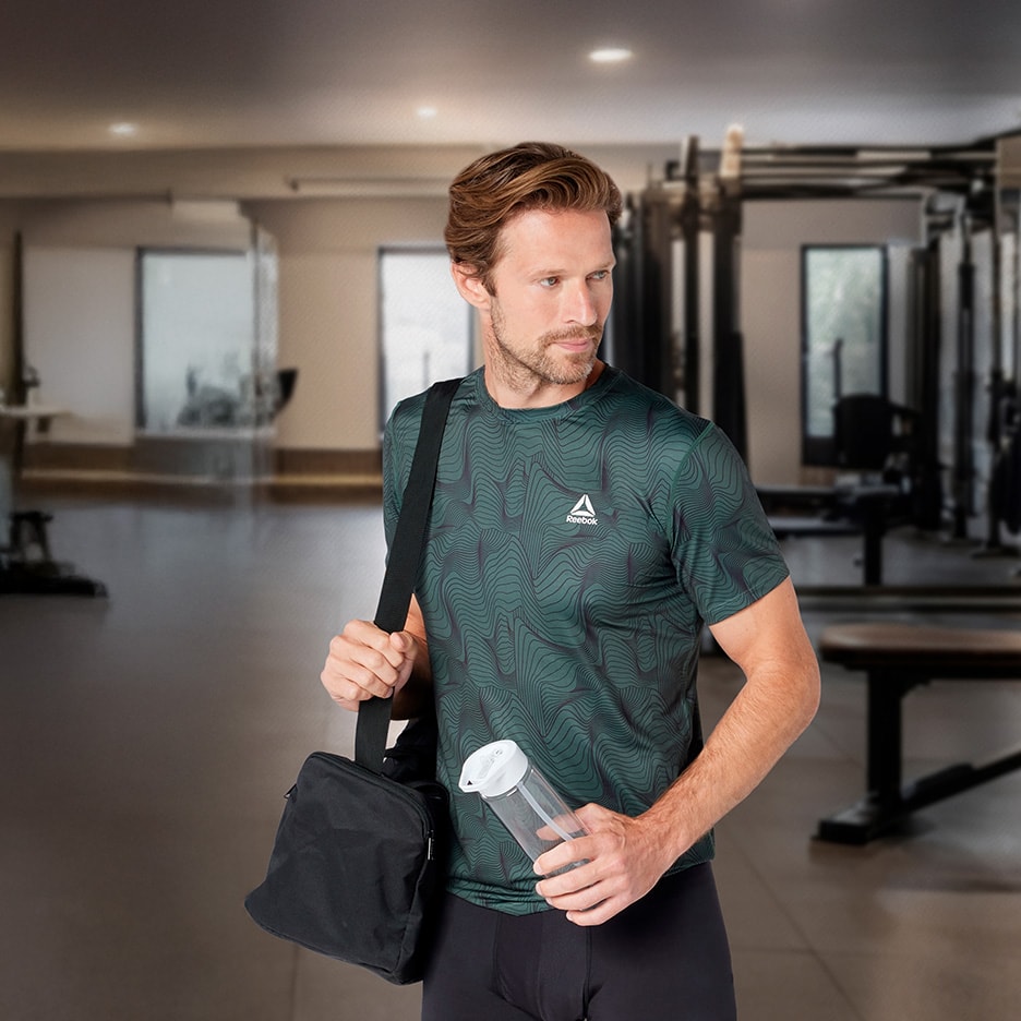 Mann in gemustertem Sport-T-Shirt und schwarzer Leggings, mit Sporttasche und Wasserflasche im Fitnessstudio.