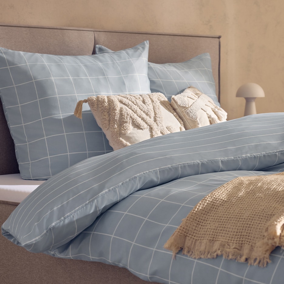 Bett mit hellblauer Bettwäsche mit Karomuster und dekorativen Kissen.