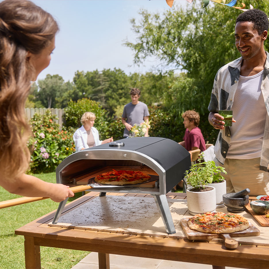 Famiglia a una festa in giardino, donna mette la pizza nel forno, uomo tiene una bevanda.
