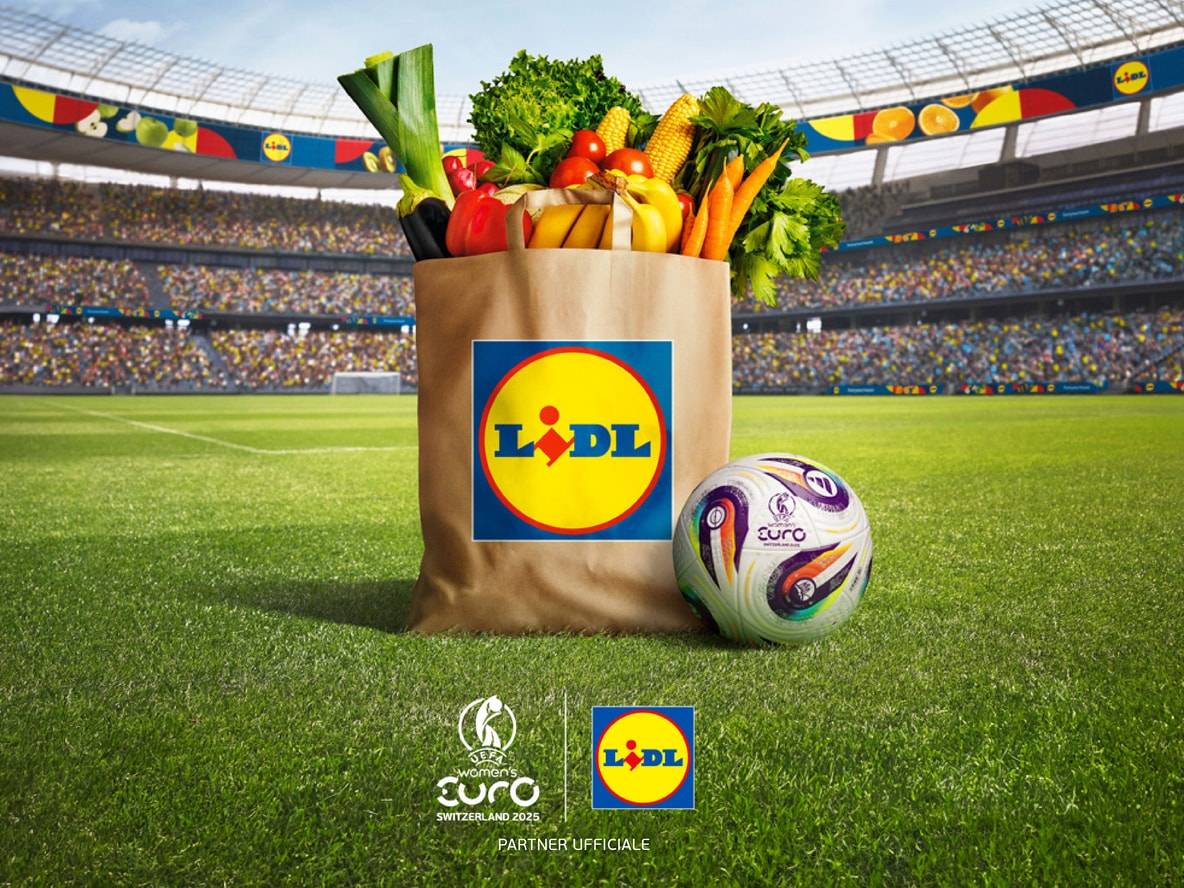 Borsa della spesa Lidl con frutta e verdura davanti a uno stadio di calcio, pallone Women's Euro 2025.
