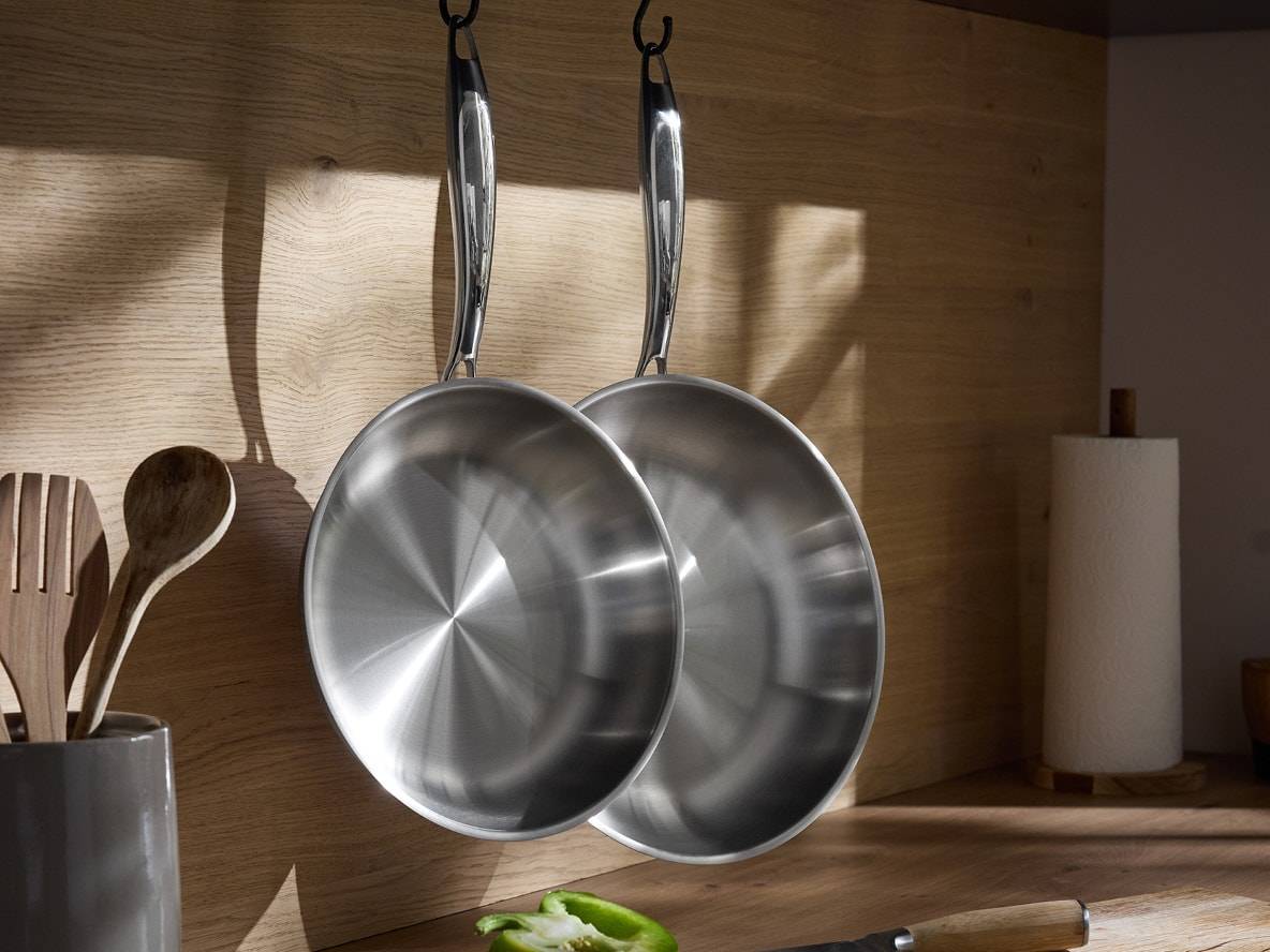 Zwei Edelstahlpfannen hängen an einer Holzwand in der Küche, daneben Holzlöffel und eine Küchenrolle.