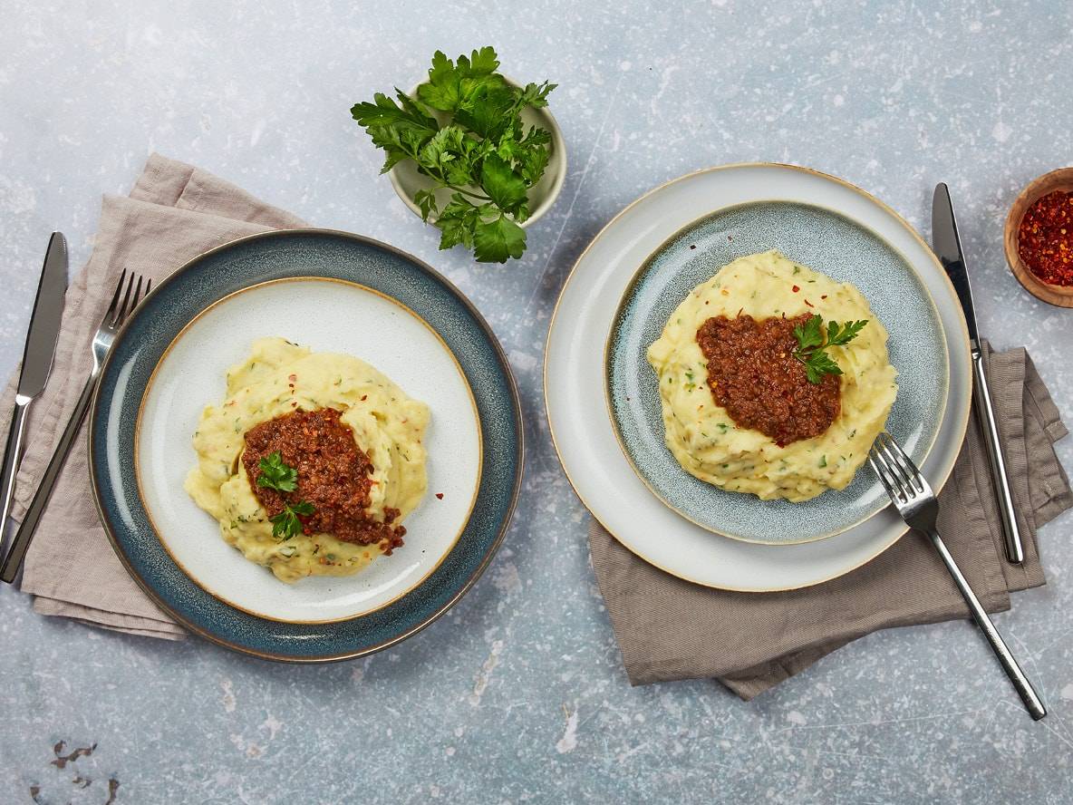 Zwei Teller Kartoffelpüree mit Hackfleischsauce und Petersilie, dazu Besteck und Gewürze.