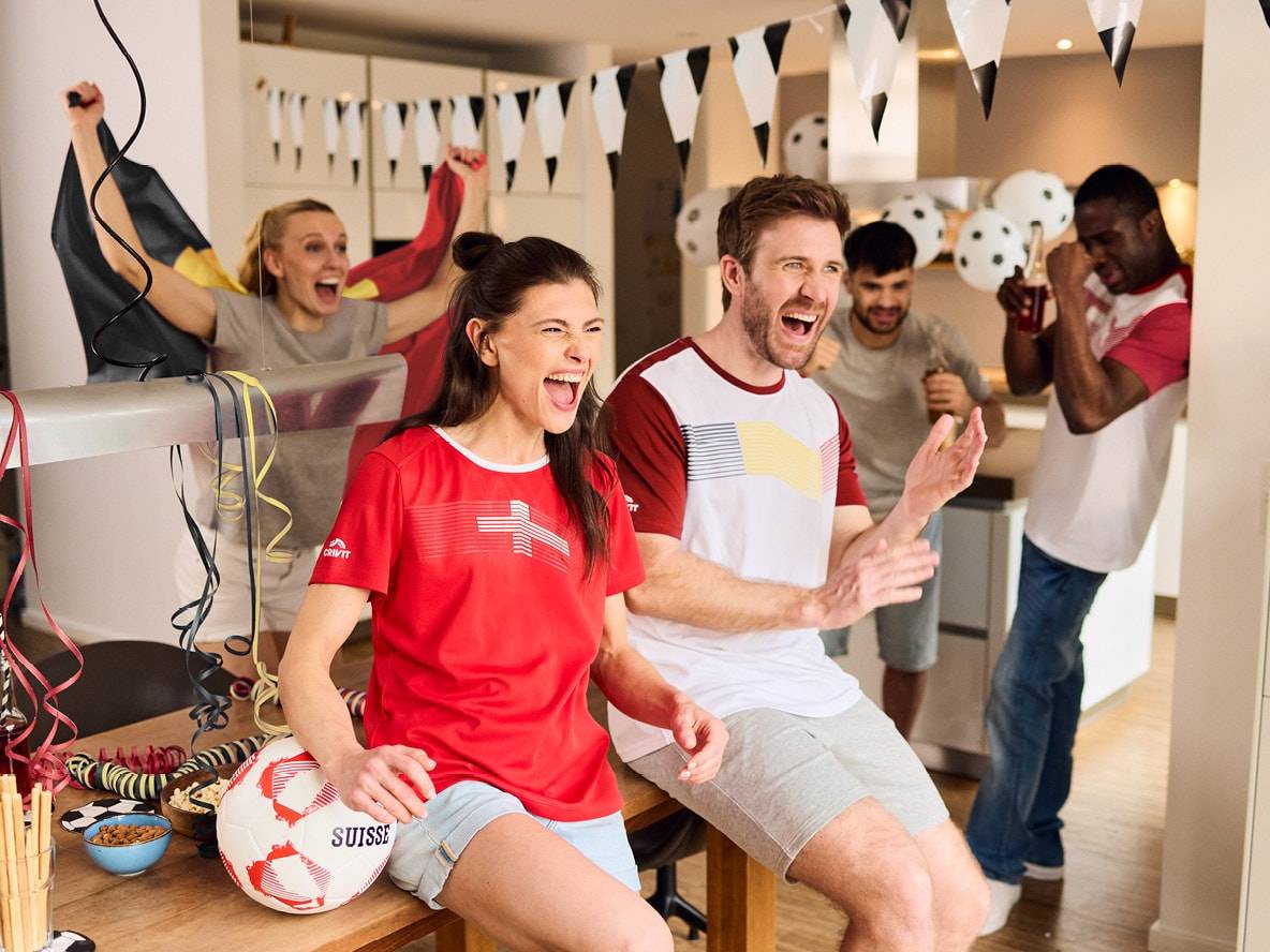 Freunde feiern mit Fußballtrikots, Flaggen und einem Fußball mit der Aufschrift „SUISSE“.