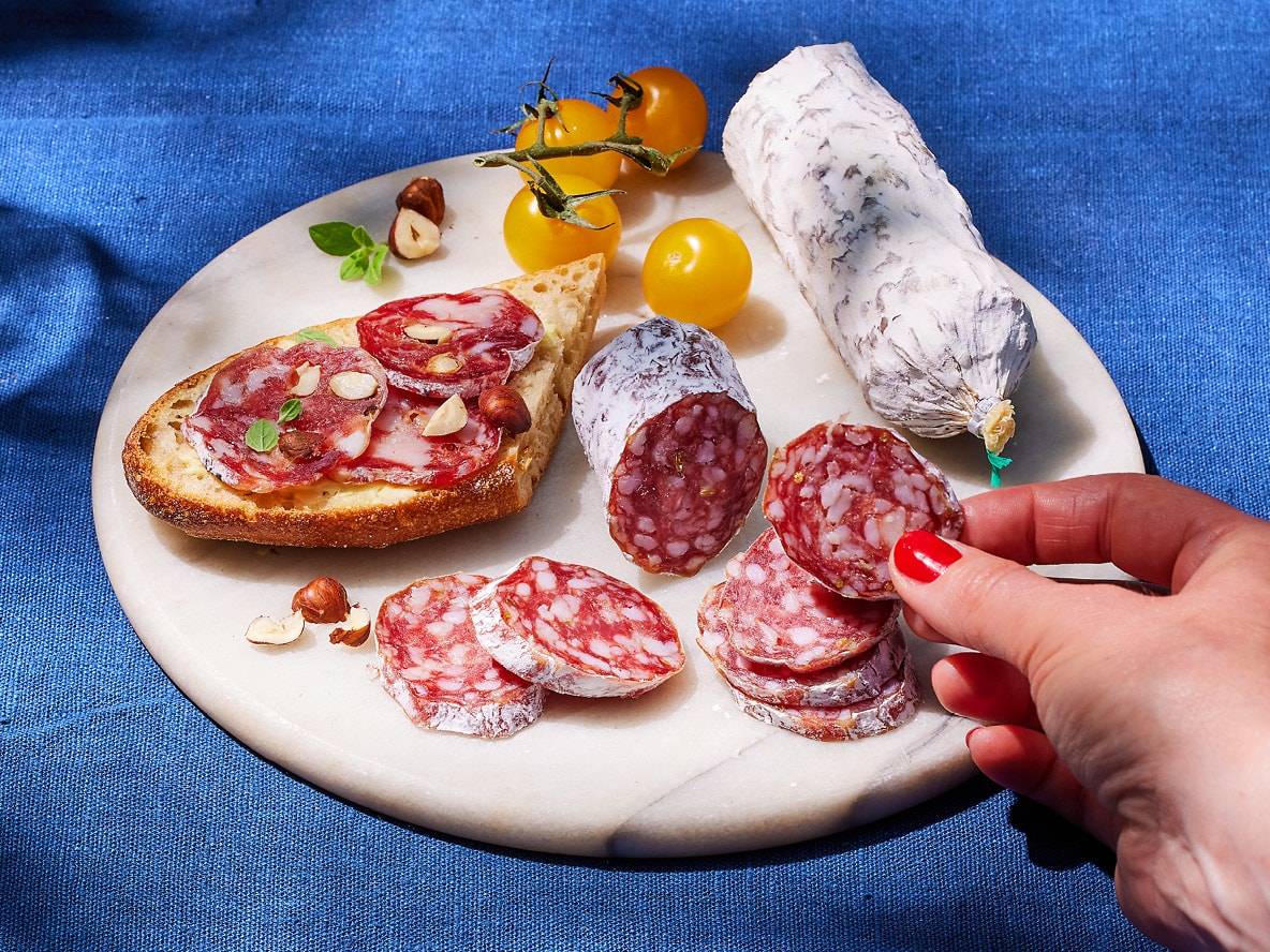 Salamischeiben und ganze Salami auf einem Marmorbrett mit Brot, Haselnüssen und Tomaten.