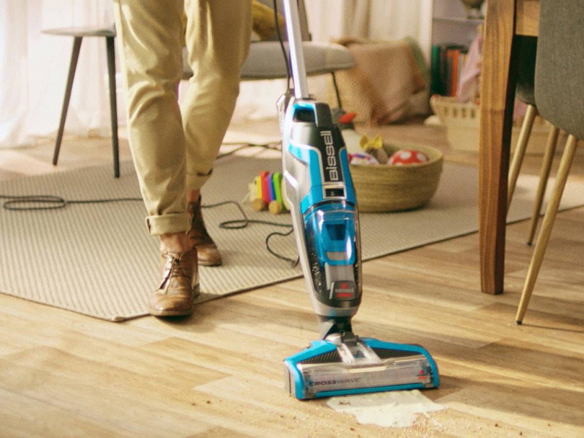 Stielstaubsauger reinigt Fleck auf Holzboden, Person steht daneben.