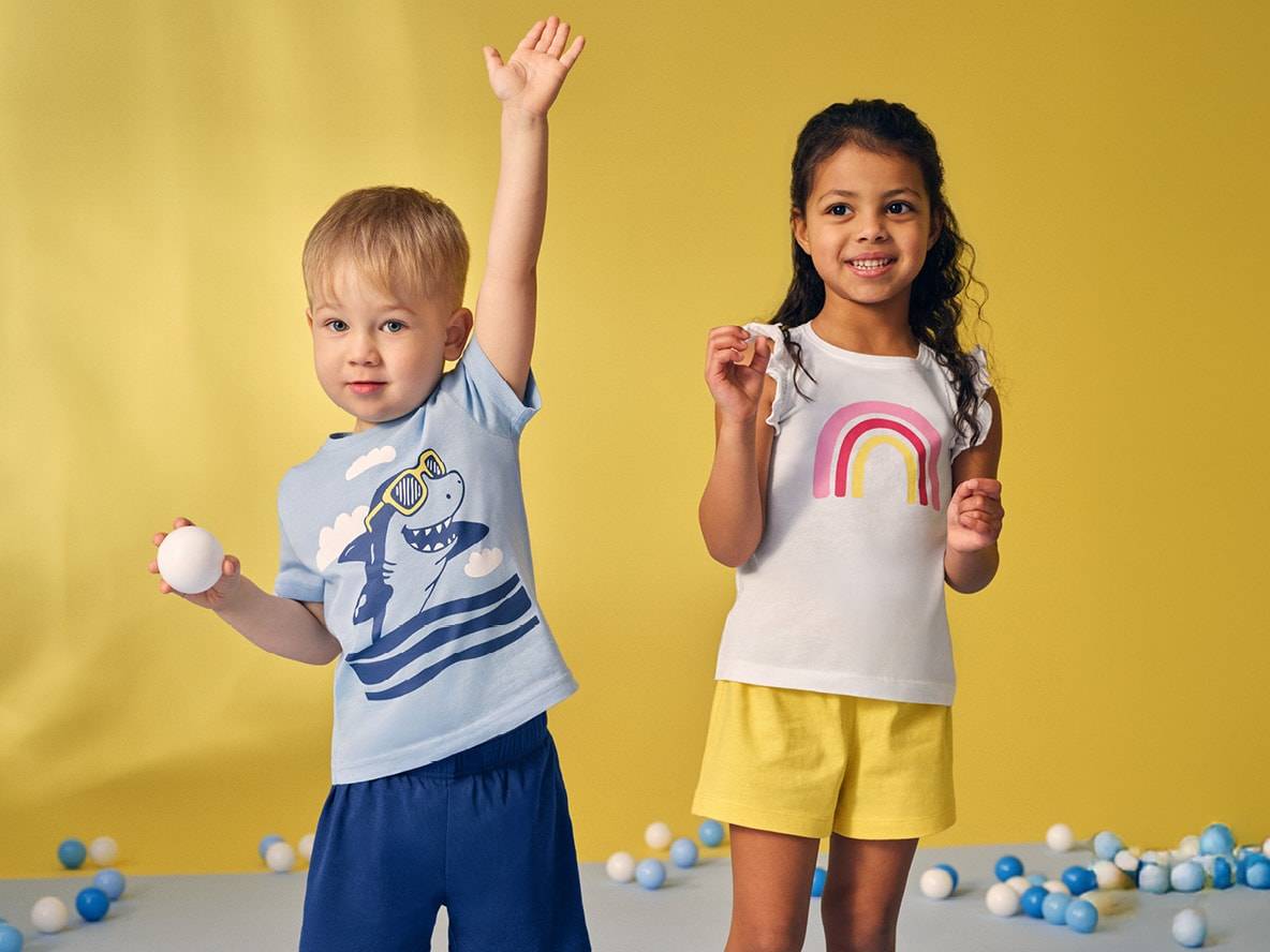 Zwei Kinder in Sommerkleidung: ein Junge in blauem Hai-T-Shirt und ein Mädchen in weißem Regenbogen-T-Shirt.