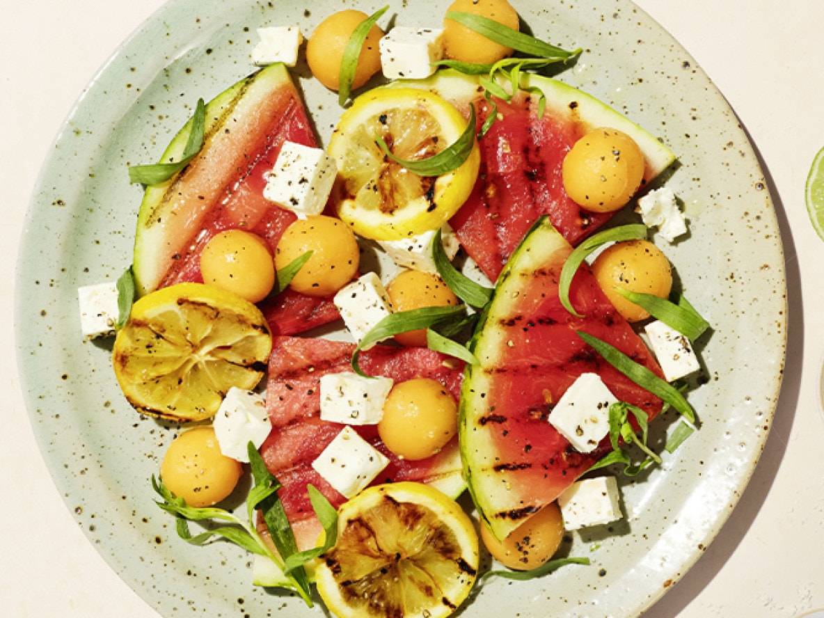 Gegrillte Wassermelone mit Feta, Melonenbällchen, Zitronen und Kräutern auf einem Teller.