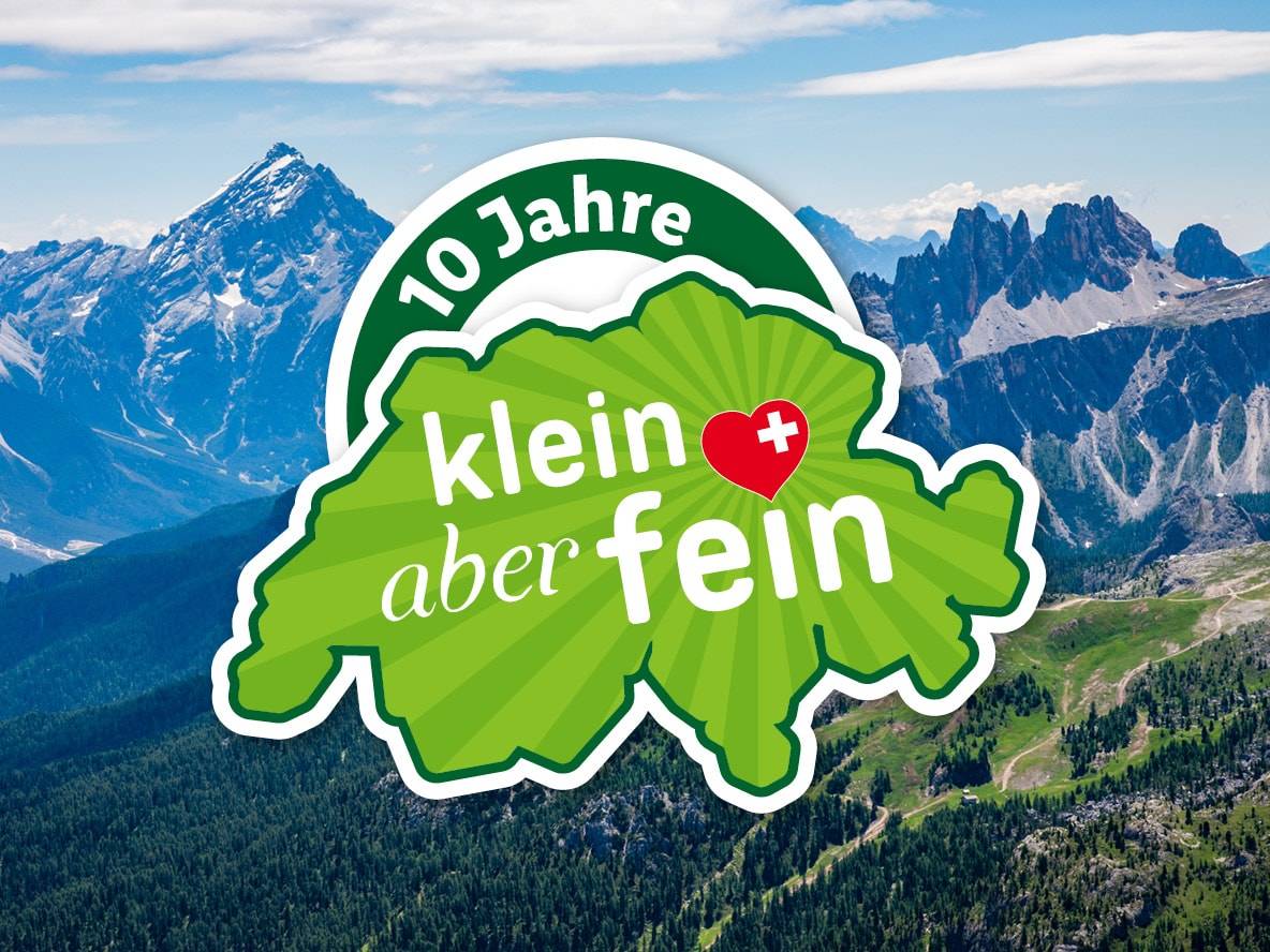 Logo '10 Jahre klein aber fein' mit Herz und Schweizer Kreuz vor Berglandschaft.