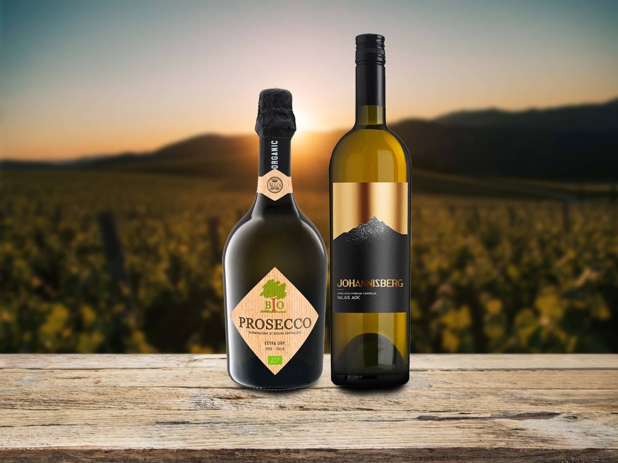 Zwei Weinflaschen, eine Prosecco und eine Weißwein, auf einem Holztisch vor einem Weinberg bei Sonnenuntergang.